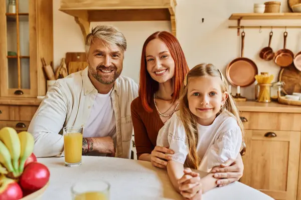 Pais alegres com filha bonita olhando para a câmera perto de frutas frescas e suco de laranja na cozinha — Fotografia de Stock