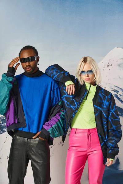 Couple multiethnique en tenues chaudes et vibrantes avec des lunettes de soleil avec toile de fond de montagne, concept d'hiver — Photo de stock