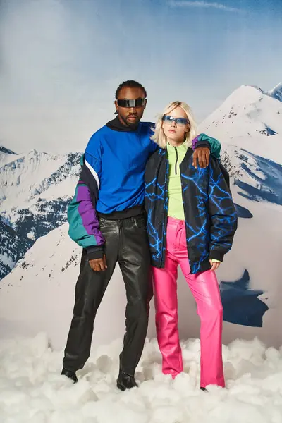 Elegante casal multirracial abraçando e posando com pano de fundo de montanha nevado, conceito de inverno — Fotografia de Stock