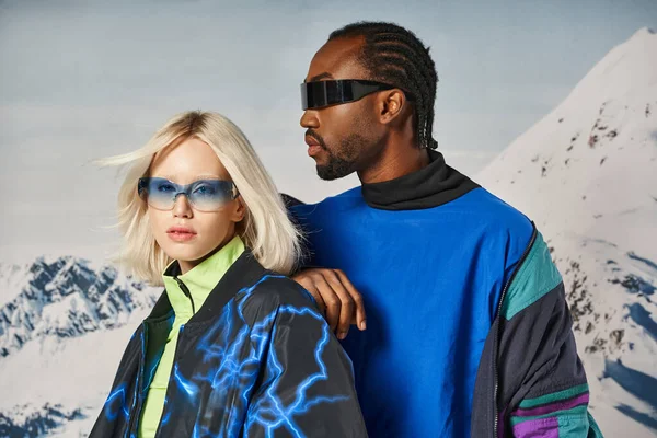 Привлекательная пара в стильных солнцезащитных очках и теплых нарядах, позирующая на снежном фоне, зимняя концепция — стоковое фото