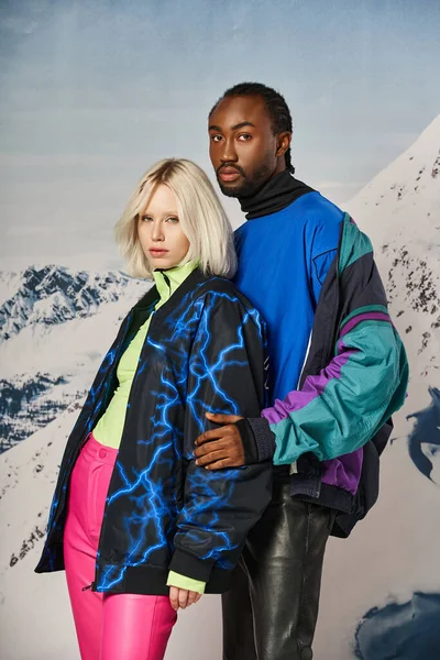 Привлекательная молодая мультикультурная пара в стильной зимней одежде обнимающая и позирующая на снежном фоне — стоковое фото