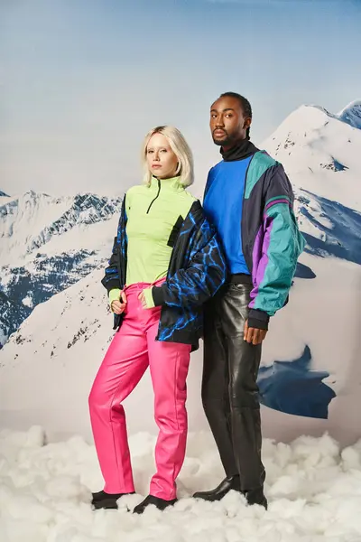 Young stylish couple in vibrant winter outfits standing on snow with mountain on background — Stock Photo