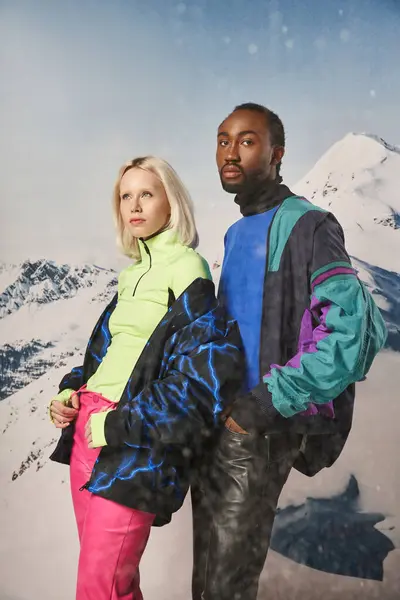 Atractiva pareja diversa en trajes cálidos con estilo con montaña nevada en el telón de fondo, concepto de invierno - foto de stock