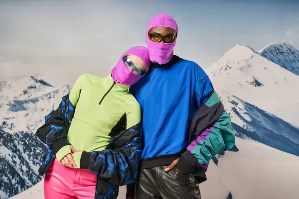 Elegante pareja diversa en pasamontañas rosadas y gafas de sol abrazando y posando juntos, concepto de invierno - foto de stock