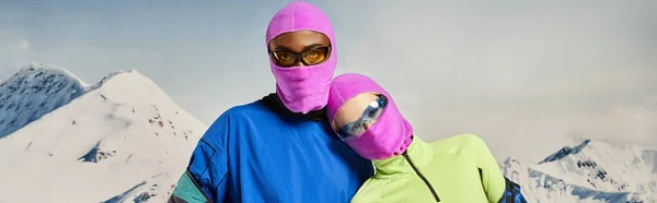 Young multiethnic couple in pink balaclavas and sunglasses hugging, winter concept, banner — Stock Photo