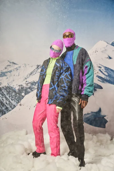 Attractive diverse couple posing together in pink balaclavas with mountain backdrop, winter concept — Stock Photo