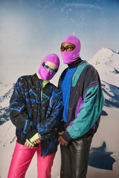 Stylish young couple in warm winter outfits and balaclavas posing together on snowy backdrop — Stock Photo