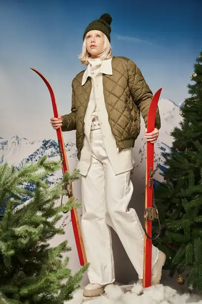 Stylish blonde woman in bobble hat and winter jacket with skis next to pine trees, winter concept — Stock Photo