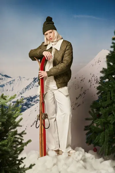 Стильная красивая женщина в теплой одежде с шапкой держа лыжи среди елок, зимняя концепция — стоковое фото
