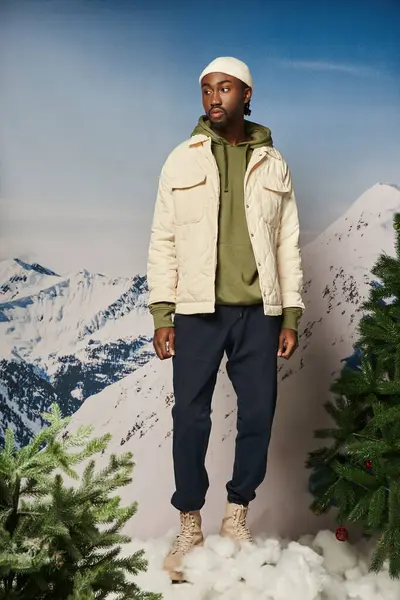 Hombre afroamericano guapo en chaqueta y gorro sombrero de pie junto a los abetos, concepto de invierno - foto de stock