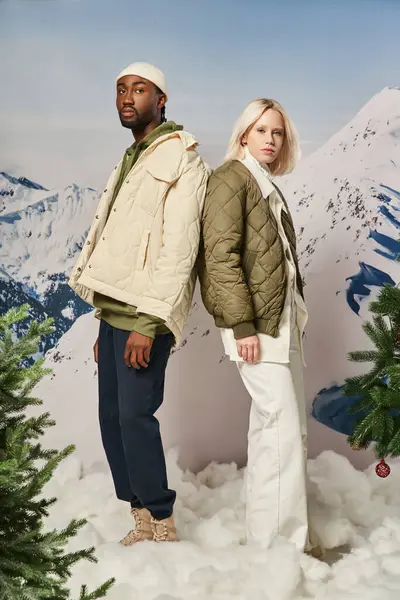 Привлекательная молодая пара в теплых нарядах, позирующая спина к спине со снежным фоном, зимняя концепция — стоковое фото