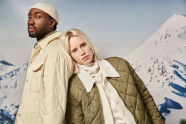 Молодая межрасовая пара в теплой одежде с горным фоном, смотрящая в камеру, зимняя концепция — стоковое фото