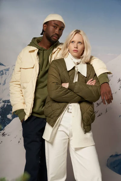 Diverse fashionable couple in warm attire posing on snowy backdrop with mountain, winter concept — Stock Photo