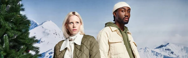 Elegante pareja multiétnica en chaquetas de moda con telón de fondo de montaña, moda de invierno, bandera - foto de stock