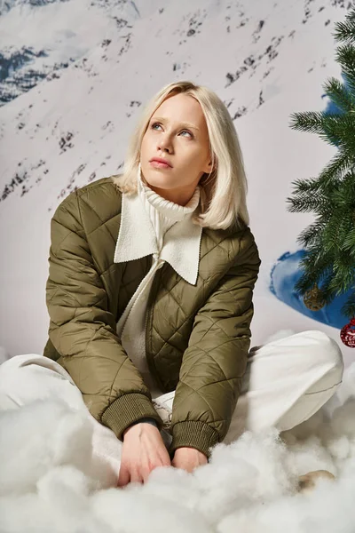 Schöne blonde Frau in warmer Winterjacke auf Schnee sitzend und verträumt wegschauend, Mode — Stockfoto