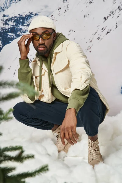 Handsome stylish man in warm modish attire squatting and touching his sunglasses, winter concept — Stock Photo