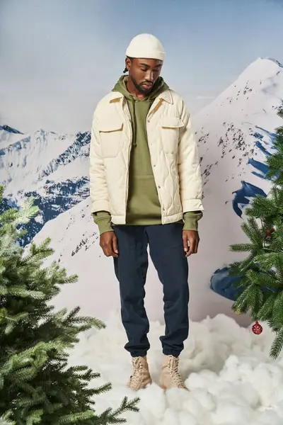 Hombre afroamericano guapo en gorro sombrero mirando hacia abajo con telón de fondo de montaña, moda de invierno - foto de stock