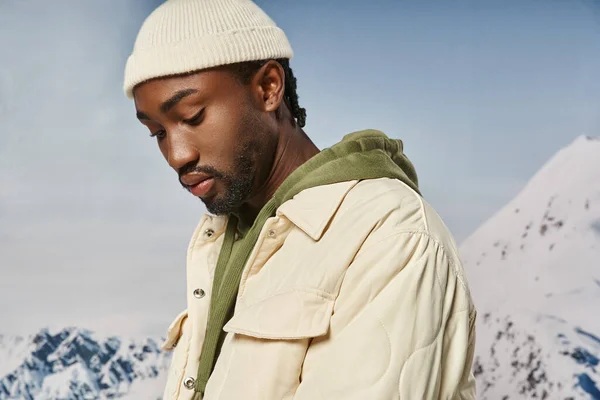 Bel homme afro-américain en veste et bonnet regardant vers le bas avec fond neigeux, mode d'hiver — Photo de stock