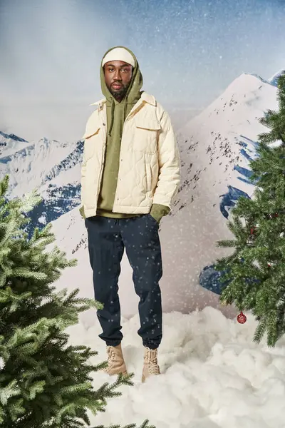 Elegante hombre de buen aspecto posando bajo la nieve con las manos en los bolsillos mirando a la cámara, invierno - foto de stock