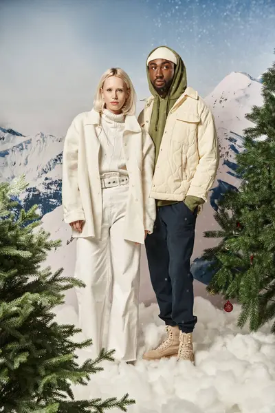 Attrayant couple diversifié posant ensemble en tenue chaude d'hiver avec fond neigeux, concept de mode — Photo de stock