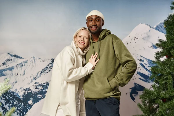 Happy blonde woman hugging with african american boyfriend with mountain backdrop, winter fashion — Stock Photo