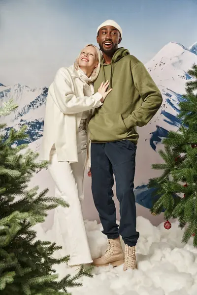 Cheerful blonde woman hugging with african american boyfriend with mountain backdrop, winter fashion — Stock Photo