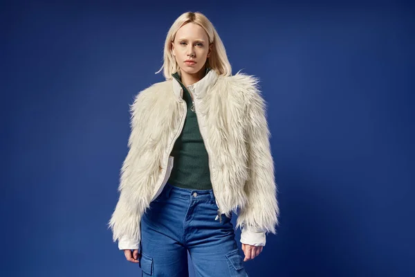 Stylish blonde woman in white faux fur jacket and denim jeans looking at camera on blue background — Stock Photo