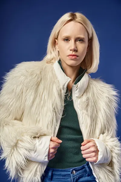 Winter fashion, stylish blonde woman in faux fur jacket and denim jeans posing on blue backdrop — Stock Photo