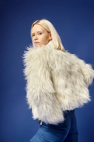 Winter attire, attractive young woman in faux fur jacket and denim jeans posing on blue backdrop — Stock Photo