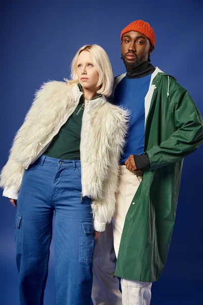 Stylish interracial couple in winter attire on blue backdrop, blonde woman and african american man — Stock Photo