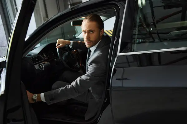 Homem de negócios bonito com barba em elegante terno inteligente atrás do volante olhando para a câmera — Fotografia de Stock