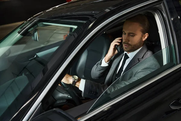 Привлекательный бородатый мужчина в элегантном костюме разговаривает по телефону за рулем, бизнес-концепция — стоковое фото