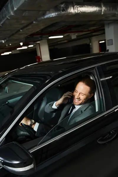 Cheerful appealing man with beard and ponytail talking by phone behind steering wheel, business — Stock Photo