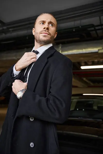 Красивий елегантний бізнесмен у стильному розумному пальто, що фіксує краватку і дивиться на парковку — стокове фото