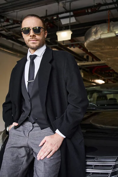 Gut aussehender schneidiger Geschäftsmann in elegantem Mantel mit Sonnenbrille und Krawatte posiert neben seinem Auto — Stockfoto