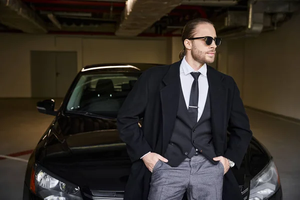 Привабливий бородатий чоловік з сонцезахисними окулярами позує біля своєї машини руками в кишенях, бізнес-концепція — стокове фото
