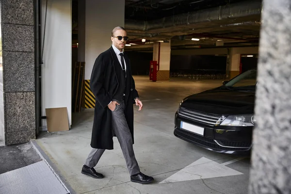 Красивый элегантный бизнесмен в черном пальто в солнечных очках, гуляющий и смотрящий в камеру — стоковое фото