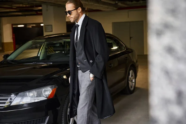 Atraente barbudo homem em casaco preto andando com a mão no bolso ao lado de seu carro, conceito de negócio — Fotografia de Stock