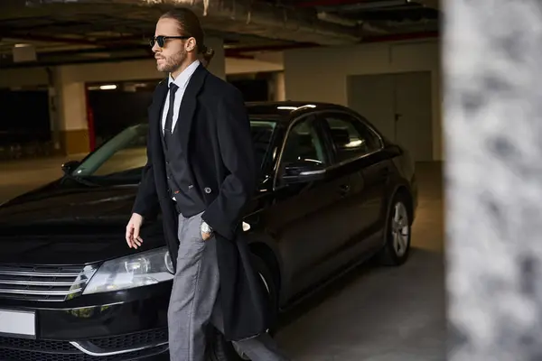 Homem de negócios bonito em casaco elegante com óculos de sol andando com a mão no bolso no estacionamento — Fotografia de Stock