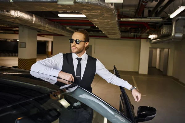 Atractivo hombre en chaleco negro con gafas de sol posando cerca de su coche y mirando hacia otro lado, concepto de negocio - foto de stock
