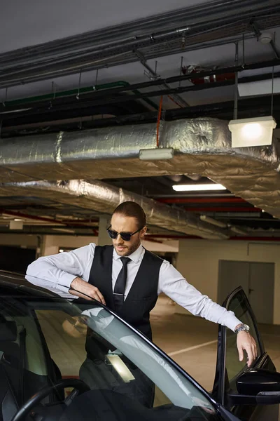 Привабливий бородатий чоловік з рудим волоссям в чорному жилеті з сонцезахисними окулярами, позує біля своєї машини, бізнес — стокове фото