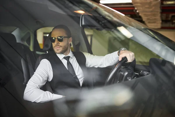 Homem barbudo atraente com óculos de sol em colete preto atrás do volante, conceito de negócio — Fotografia de Stock
