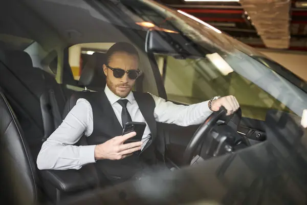 Schöner charmanter Geschäftsmann in schwarzer Weste mit Sonnenbrille, der hinter dem Lenkrad auf sein Handy blickt — Stockfoto