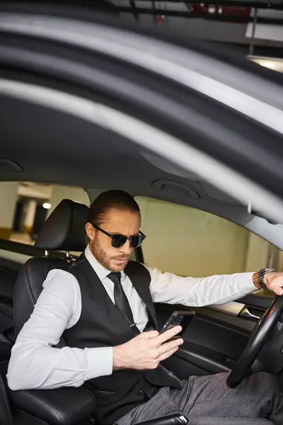 Appealing red haired man with beard and sunglasses looking at phone behind steering wheel, business — Stock Photo