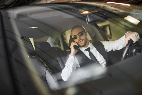 Attractive bearded man with ponytail and sunglasses talking by phone behind steering wheel, business — Stock Photo