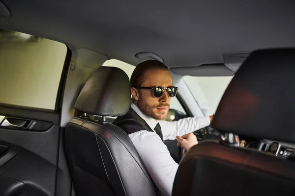 Gut aussehender bärtiger Geschäftsmann mit Sonnenbrille blickt hinter dem Lenkrad in die Kamera — Stockfoto