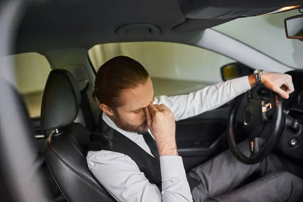 Tired bearded businessman with ponytail in black vest sitting behind steering wheel of his car — Stock Photo