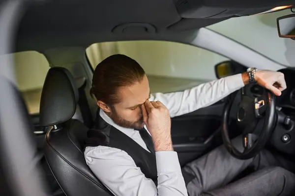 Müder bärtiger Geschäftsmann mit Pferdeschwanz in schwarzer Weste sitzt am Steuer seines Autos — Stockfoto