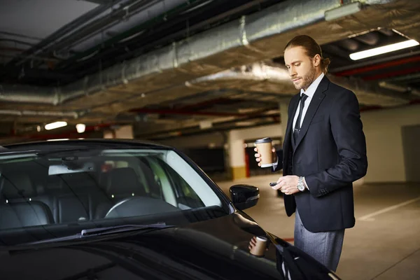 Hombre elegante de buen aspecto en traje negro con corbata sosteniendo taza de café al lado de su coche, concepto de negocio - foto de stock