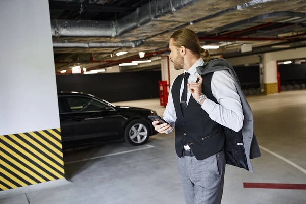 Гарний вигляд бородатого чоловіка в чорному жилеті, що тримає мобільний телефон і дивиться на його машину, бізнес-концепція — стокове фото